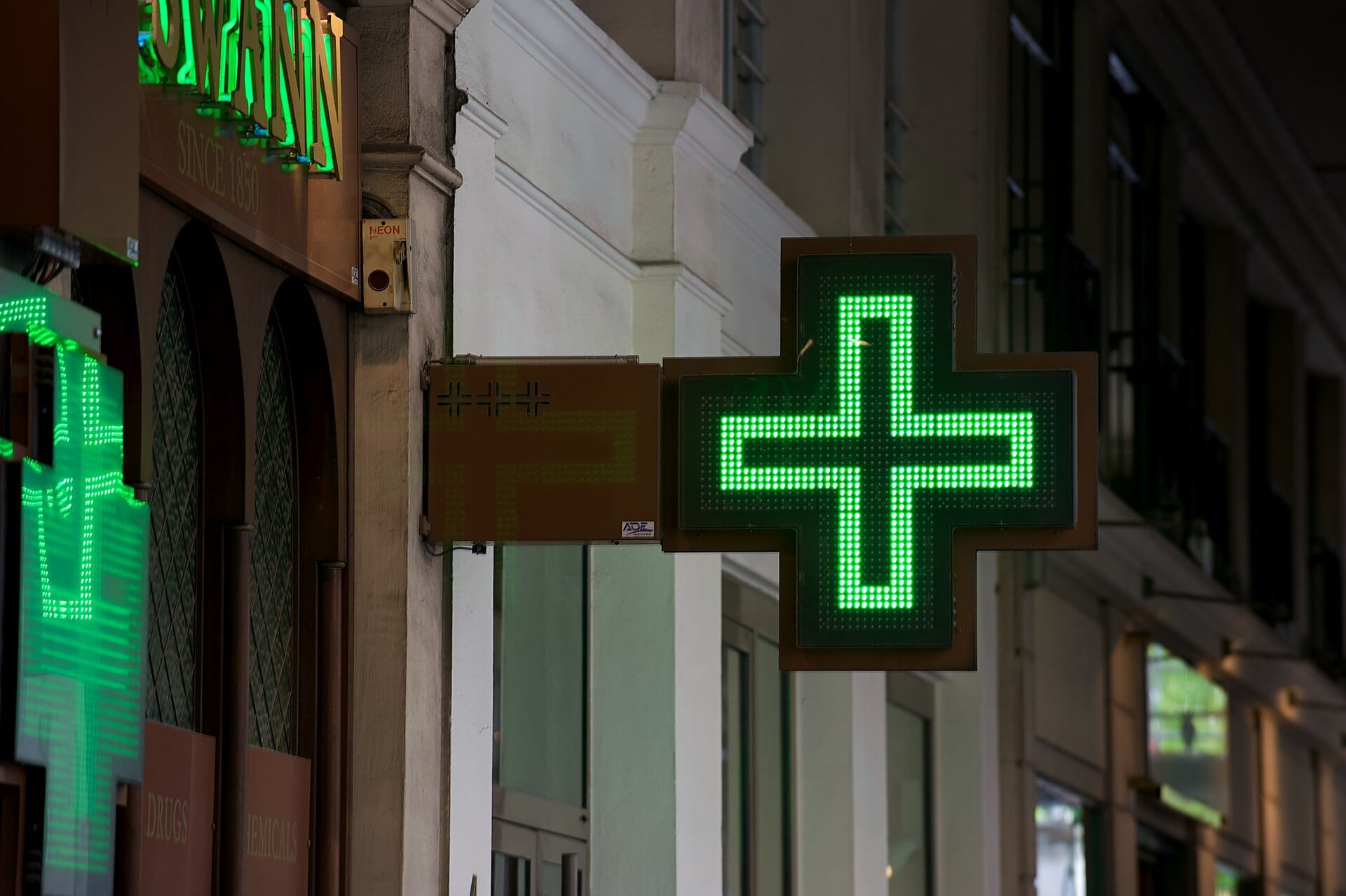 A lit green pharmacy cross hanging over a street