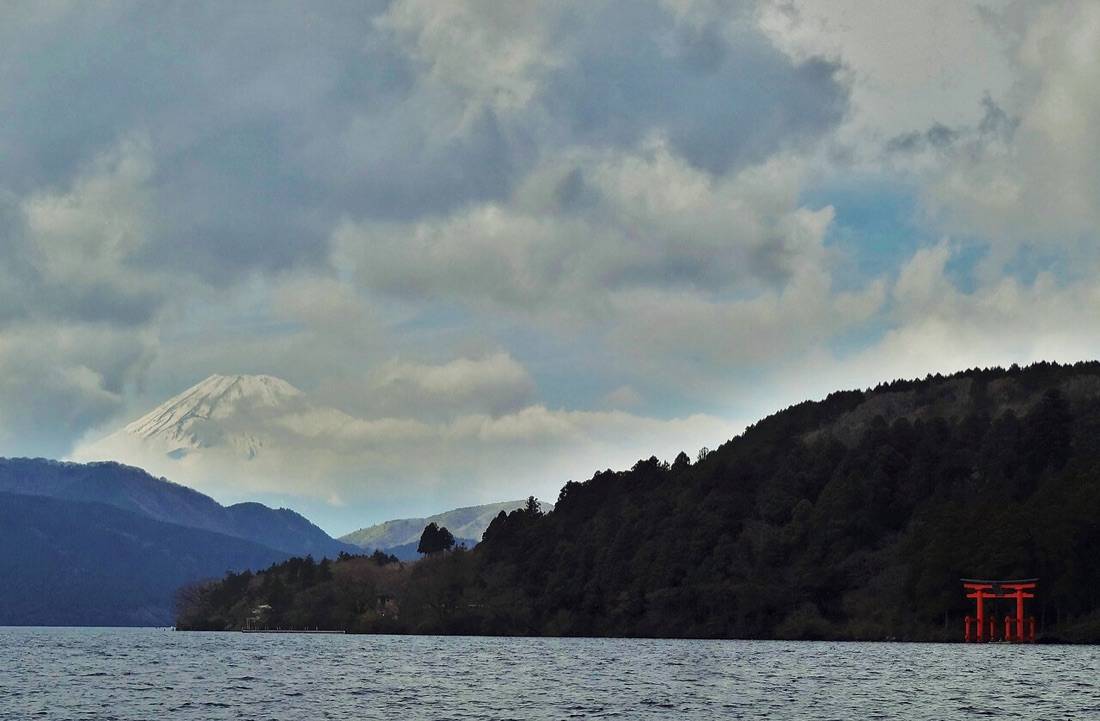 Snowcapped Mount Fuji above the wooded shore of Lake Ashi, with the red torii of Hakone Shrine at the water’s edge