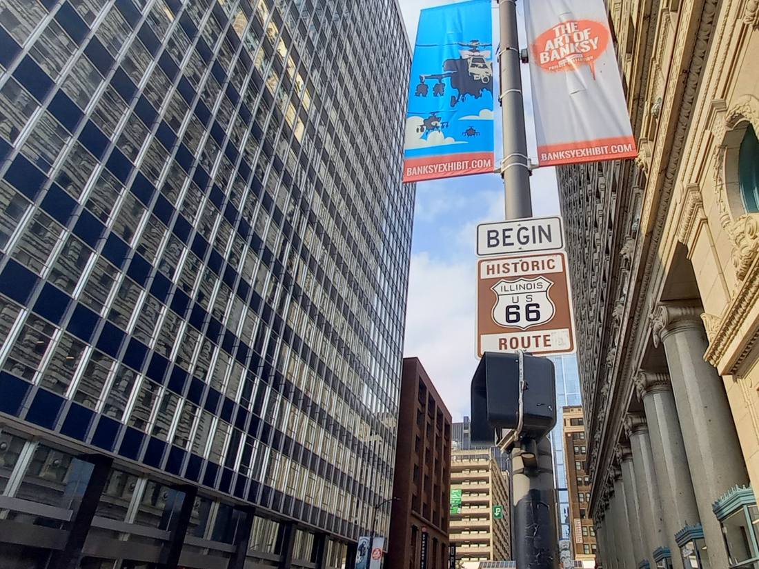 A brown Begin Historic Route 66 sign on a lamppost between downtown Chicago towers