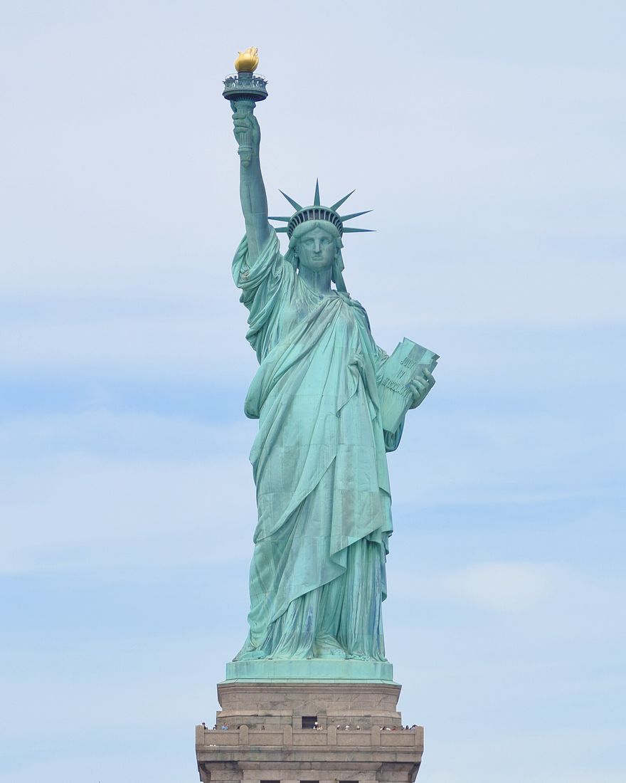 The green copper Statue of Liberty holding up her torch against a pale sky