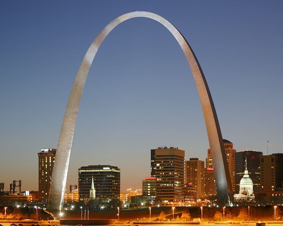 The steel Gateway Arch lit up at dusk over the St. Louis skyline and the Mississippi